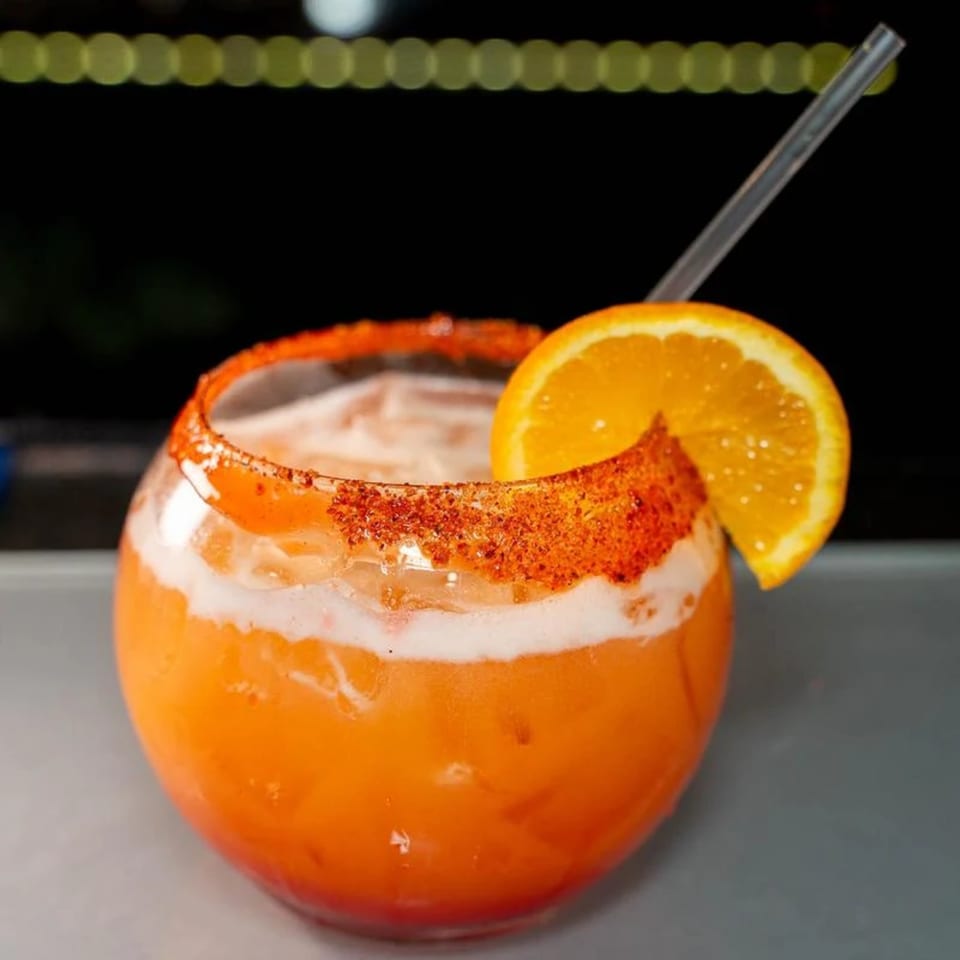 A vibrant orange Mexican cocktail with a chili-lime rim, orange slice, and straw.