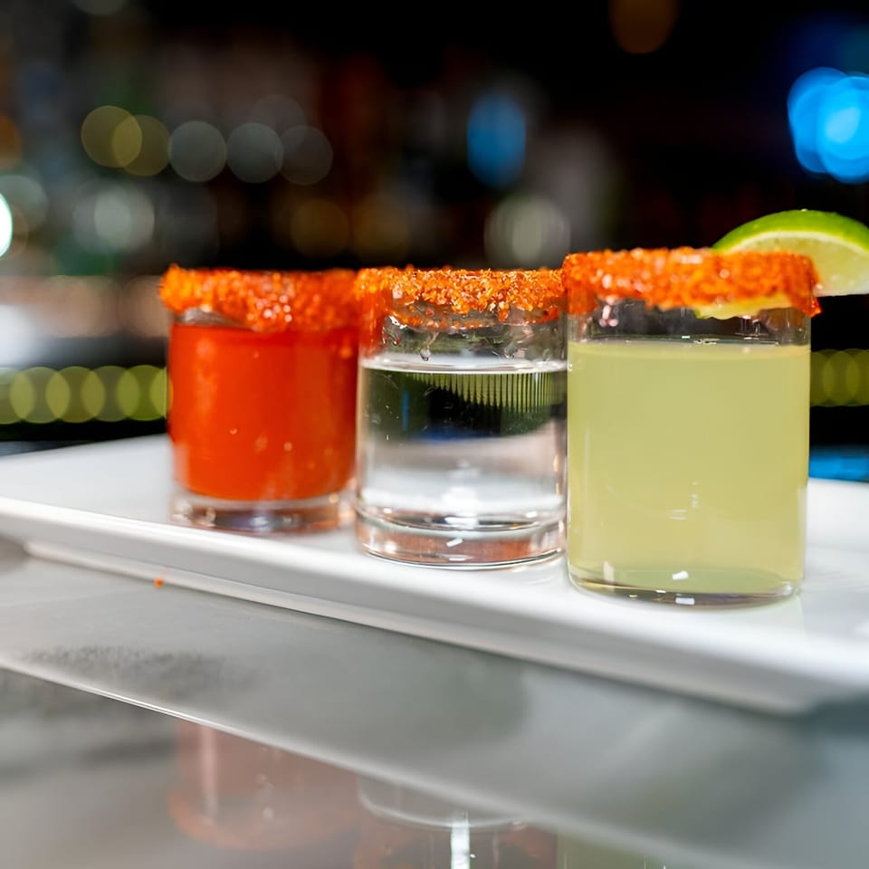 Three Mexican-style shots with chili-lime rims, one red, one clear, and one yellow with a lime wedge.