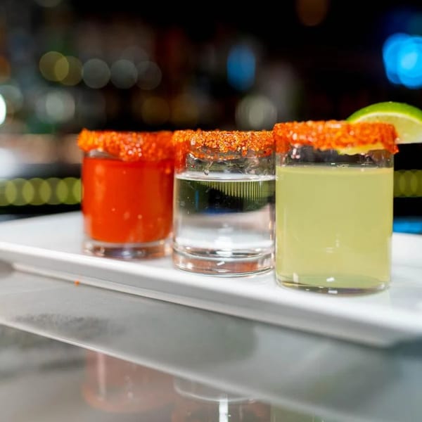 Three Mexican-style shots with chili-lime rims, one red, one clear, and one green with a lime wedge.