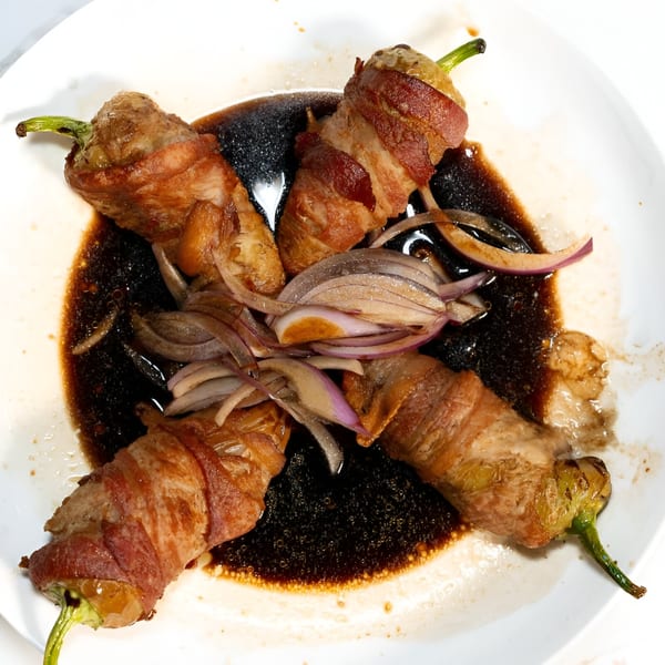 Four bacon-wrapped jalapeño poppers with sliced red onions in a dark sauce on a white plate.