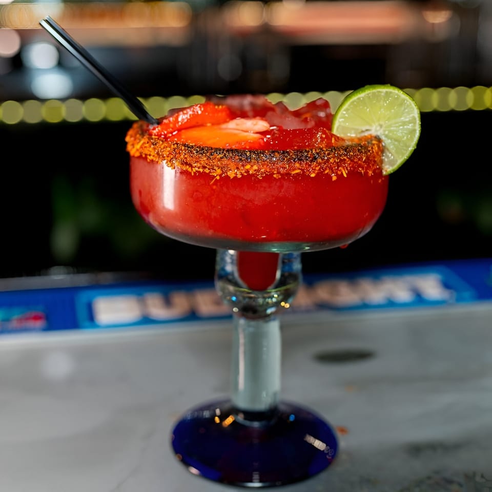 A vibrant red Mexican cocktail, garnished with chili salt, lime, and strawberries, on a bar.